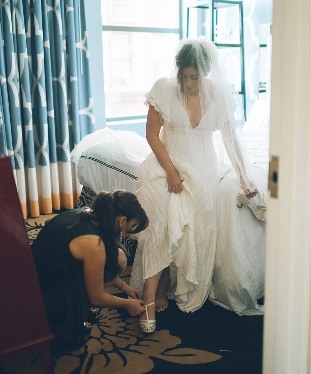 A bridesmaid kneeling to fasten Hannah's LunaSol Caged Swing heels while she gets ready